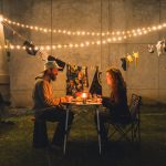 A Man And A Woman Sitting At A Table Under String Lights, Enjoying Dinner While Clothes Hang On A Line In The Background
