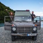 Eine Frau Und Ein Mann Lehnen Sich Froehlich Aus Einem Puch Gelaendewagen, Im Hintergrund Ein Fluss Und Wald