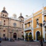 Eine platz vpller menschen mit einer alten sandsteinfarbenen kirche und einem gelb weissen kolonialen gebäude